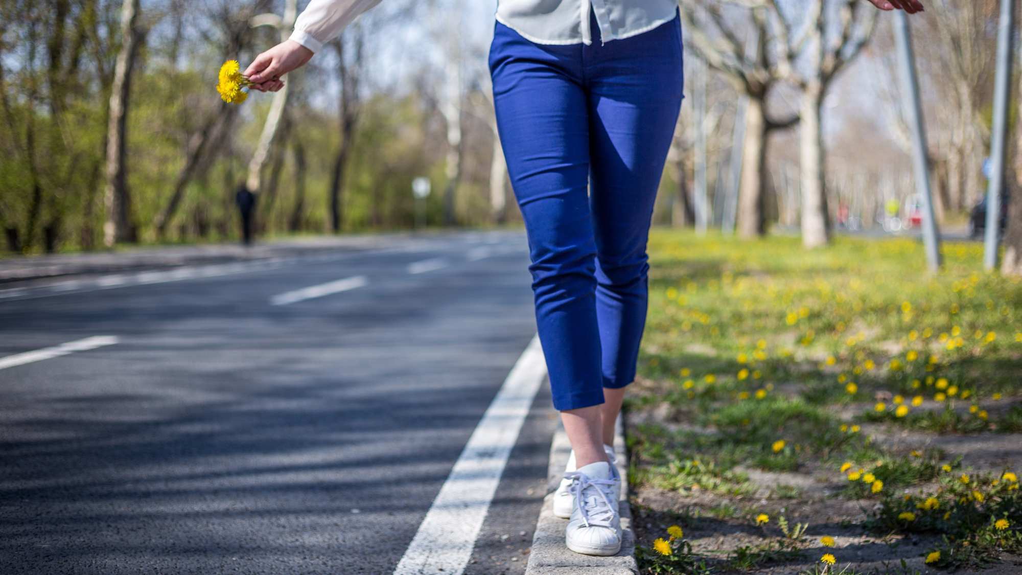 An image of a woman from the waist down wearing blue pants and a white shirt walking and balancing on the white line on the side of a road. She has yellow flowers in her hand.