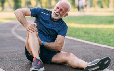 In image of an man with white hair wearing workout clothing, sitting on a track and clutching his knee in pain.
