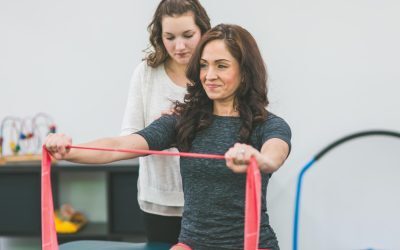 An image of two Caucasian women, one is doing Physical Therapy exercises for her shoulder, the other is her therapist and is standing behind her while she exercises.