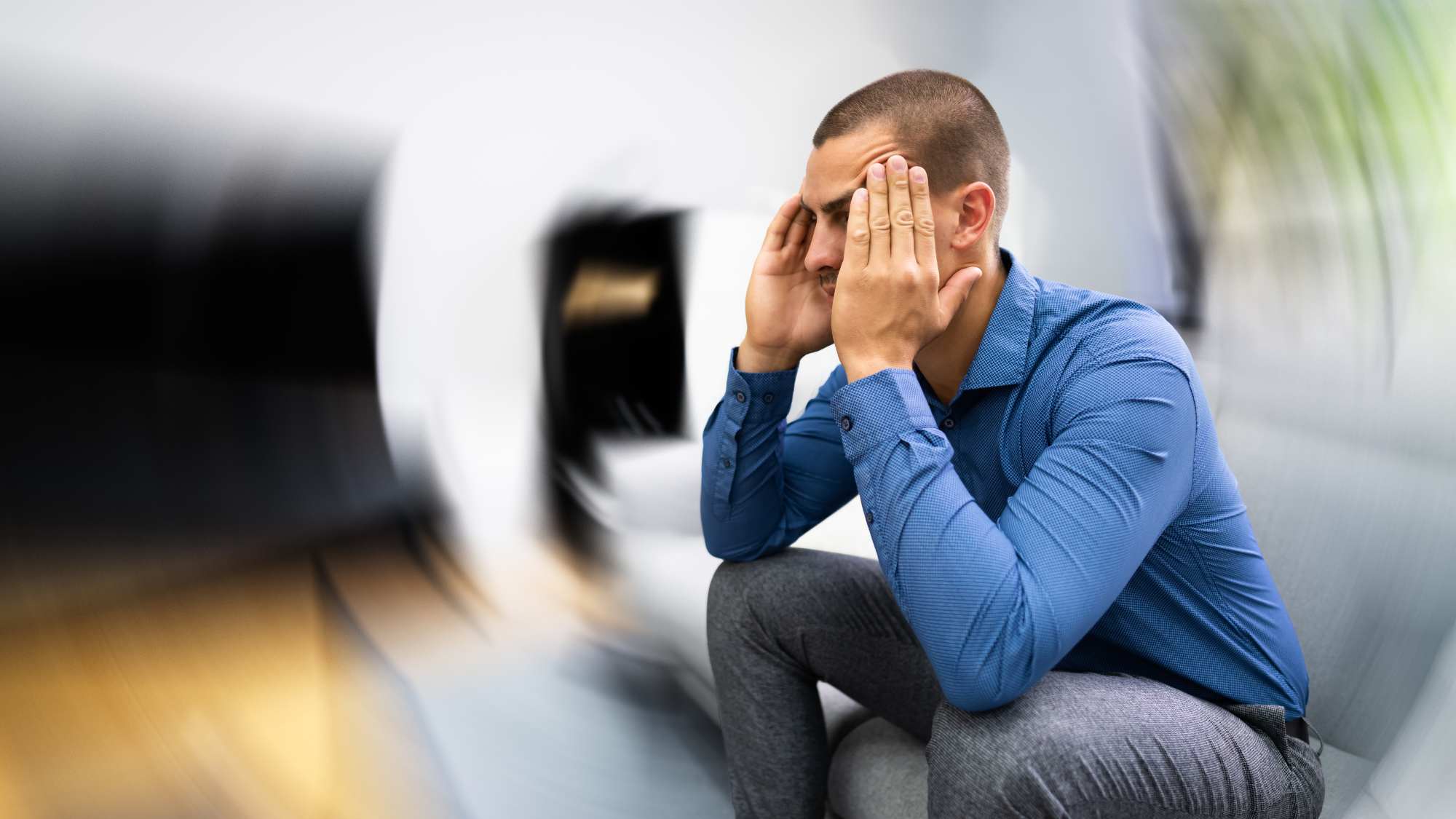An image of a man sitting on a couch holding his head in his hands. There are blur lines around the outside of the man, indicating that he feels dizzy or off balance.